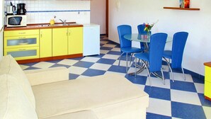 Una cocina compacta con armarios amarillos, un refrigerador blanco y un microondas. Hay un comedor con una mesa de vidrio y sillas azules.