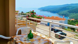 Un balcón con una mesa dispuesta para dos, con vistas a un paisaje costero que incluye el mar y colinas.