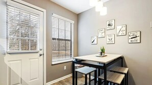 Una cocina con una puerta blanca, dos ventanas con persianas, una mesa con sillas y decoración en la pared.