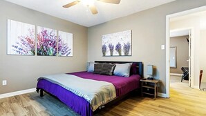 Un dormitorio con cama, mesita de noche, ventilador de techo y dos cuadros florales colgados en la pared.