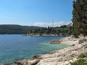 A coastal landscape with rocky terrain, clear blue water, and a distant hill with buildings. - Nice apartment for 4 people with pool, A/C, WIFI, TV and terrace (Labin)
