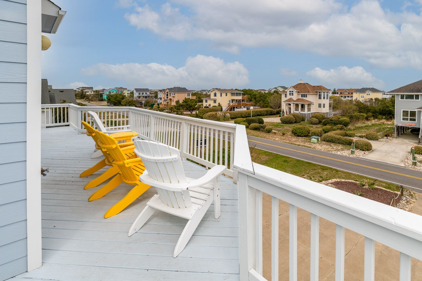 Un balcon agrémenté de chaises Adirondack blanches et jaunes, avec vue sur un quartier résidentiel, ses maisons et une route.