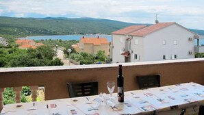 A balcony with a table set for dining, overlooking a lake and buildings in a hilly area. - Nice apartment for 4 guests with WIFI, A/C, TV, balcony and pets allowed (Novi Vinodolski)