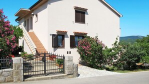 Une maison à deux étages, avec un toit en tuiles, une cheminée et une entrée fermée par un portail.