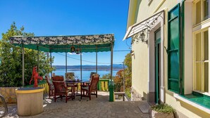 Eine Terrasse mit Tisch und Stühlen, ein überdachter Bereich mit Meerblick und ein Gebäude mit grünen Fensterläden.