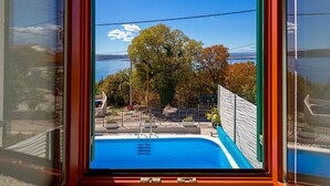 Blick aus einem Fenster auf ein Schwimmbecken, mit blauem Himmel und Bäumen im Hintergrund.