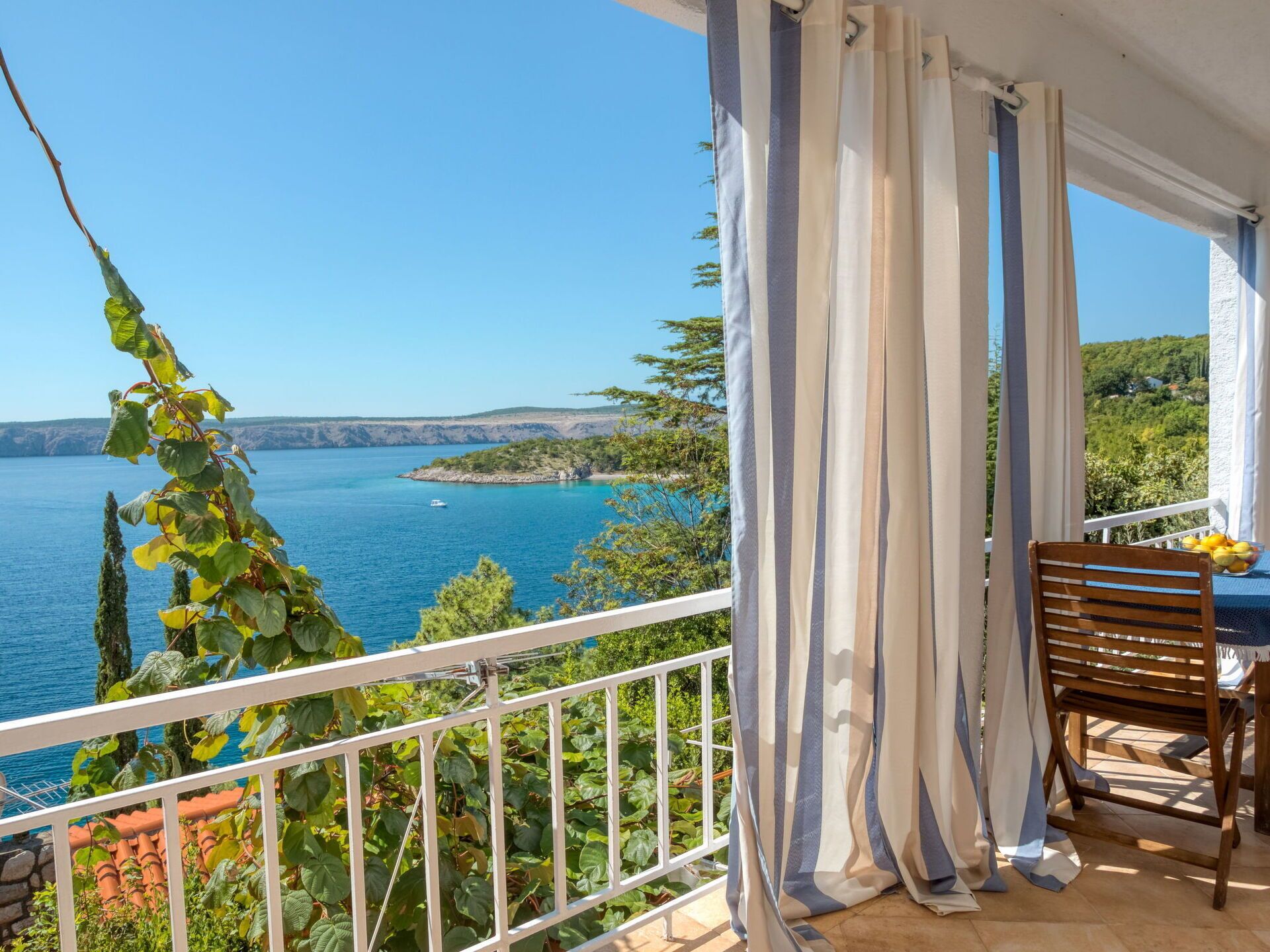 A balcony with a view of a body of water, a chair, and a table with fruit.