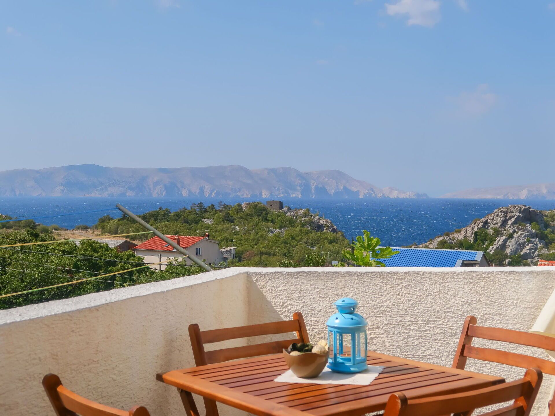 A balcony with a table, chairs, a lantern, and a view of the sea and mountains.