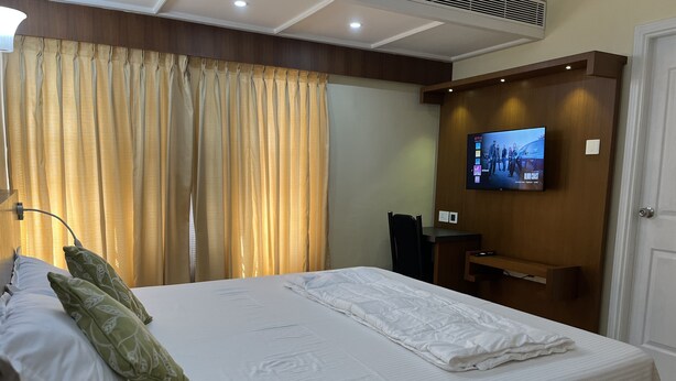 A hotel room with a bed, curtains, a television on the wall, and a desk. - Studio Apartment Suite with kitchenette (THIRUVANANTHAPURAM)