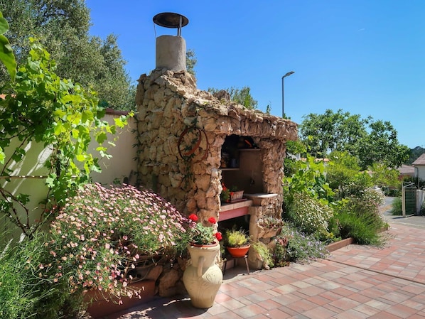 A stone-built outdoor kitchen with a chimney, surrounded by potted plants and flowers.