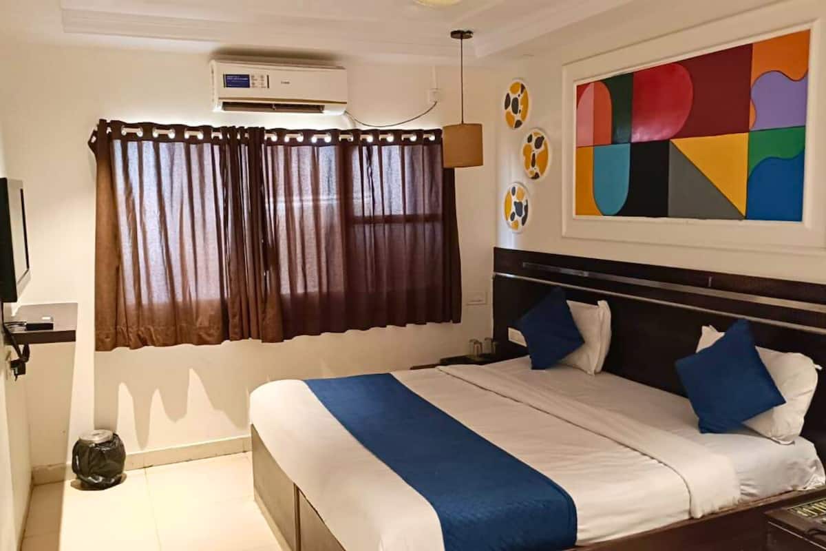 A hotel room with a bed, curtains, a wall-mounted air conditioner, a ceiling fan, and a colorful abstract painting.