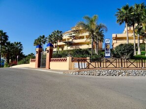 Una entrada con portón, techo azul, palmeras y un edificio de varios pisos con balcones.