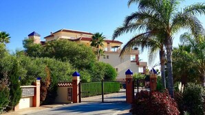 A gated entrance to a building with a red-tiled roof, surrounded by lush greenery and palm trees.