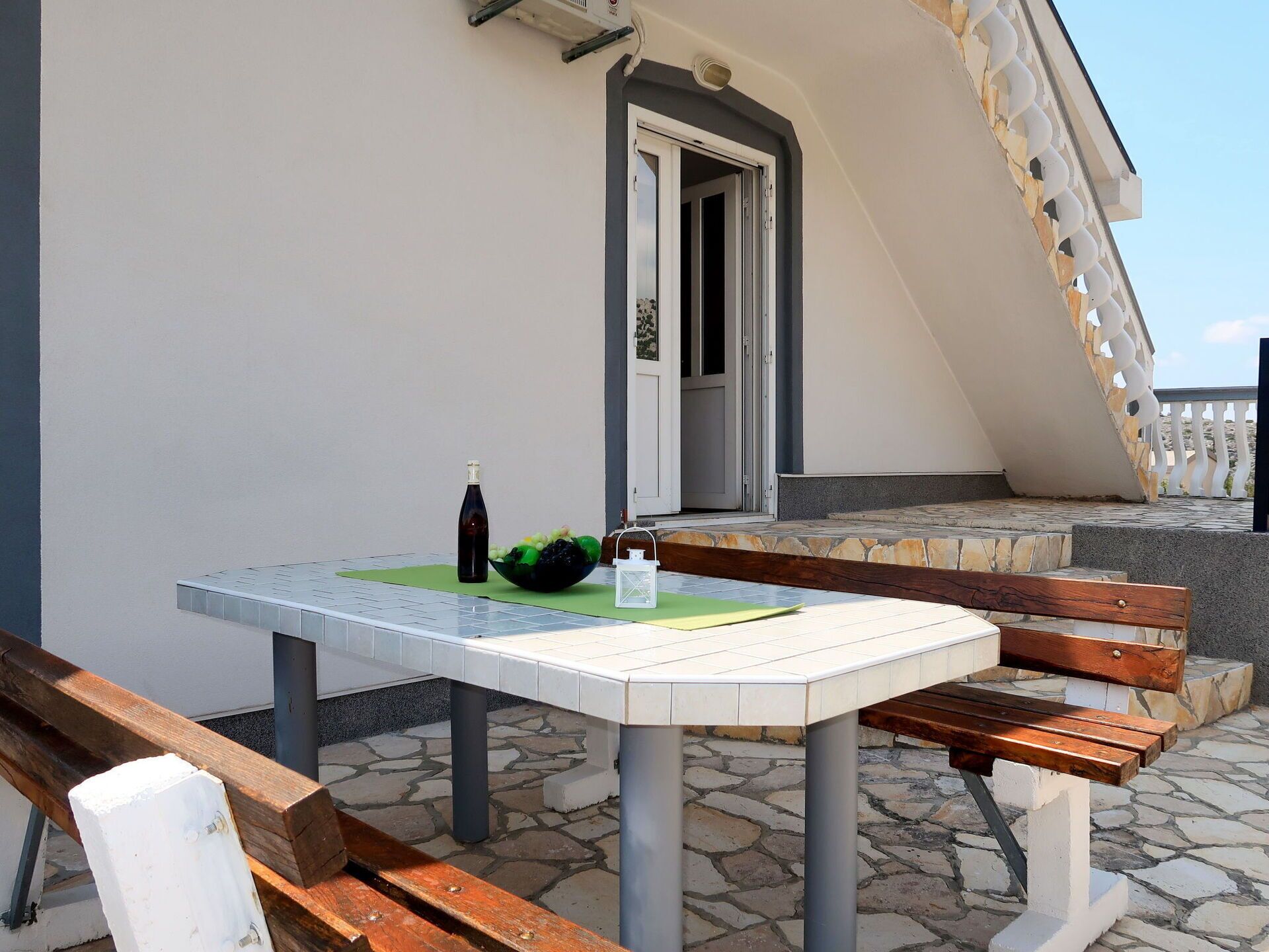 Une table avec une bouteille et un saladier de fruits, un banc en bois et un sol carrelé en pierre.