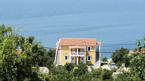 A two-story yellow building with a red-tiled roof, located near a body of water with a clear view of the sea and distant mountains.
