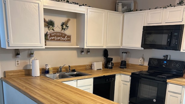 Una cocina con armarios blancos, encimera de madera, estufa negra y microondas. Hay dos taburetes de bar y una cafetera.