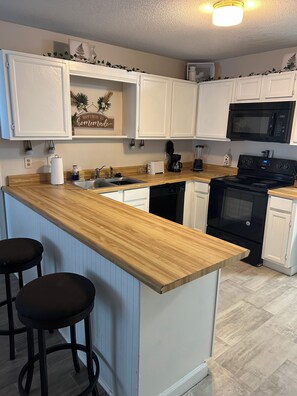 Una cocina con armarios blancos, encimera de madera, estufa negra y microondas. Hay dos taburetes de bar y una cafetera.