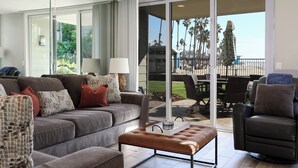 A modern living room with a grey sofa, brown ottoman, and a black chair. A glass door leads to a balcony with outdoor furniture and palm trees. - Your Own Slice of Paradise! Patio & Grass Just Steps from the Sand! (Oceanside)