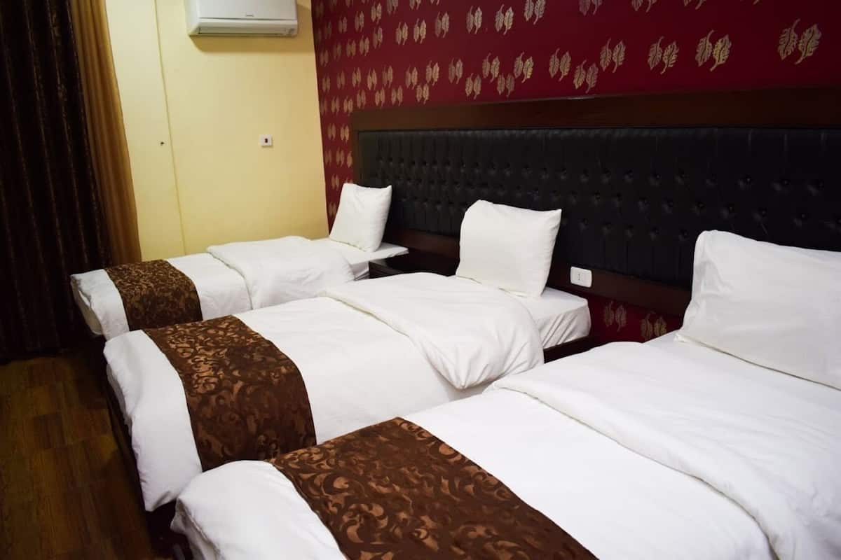 Habitación de hotel con tres camas, cada una con sábanas blancas y almohadones marrones. La habitación tiene un papel tapiz rojo con estampado y un cabecero oscuro.