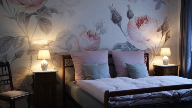 Une chambre à coucher avec un papier peint à motifs floraux, un lit avec des oreillers, deux tables de chevet avec des lampes et une chaise.