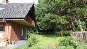 Une maison d’habitation avec un toit en pente, une façade en briques et un petit jardin devant, agrémenté de diverses plantes et d’une clôture en bois.