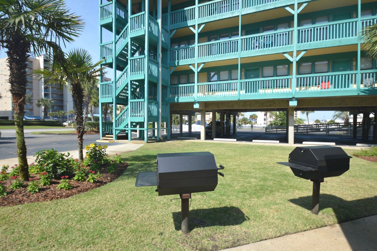 A building with a green exterior and multiple balconies. There are two barbecue grills on the lawn in front of the building.
