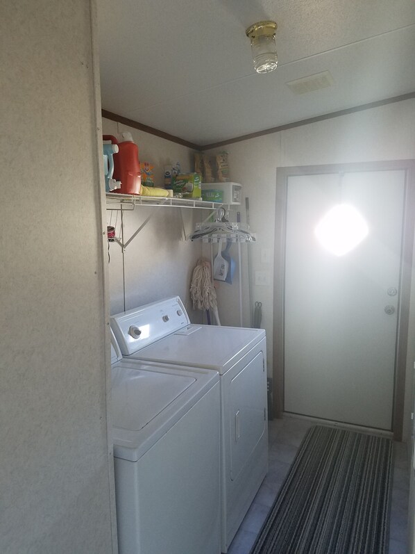 Un lavadero con lavadora y secadora, un estante con productos de limpieza, y una puerta con una luz encima.