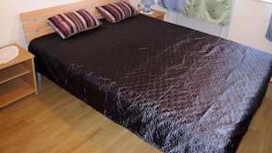 Een enkel bed met een donkere quilt, een houten hoofdbord, twee gestreepte kussens, een nachtkastje met een lamp en een raam met gordijnen.