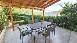 Un patio cubierto con mesa y sillas, rodeado de vegetación y vistas al mar.