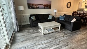 Un soggiorno con pavimento in legno, due divani neri, un tavolino da caffè bianco e un quadro raffigurante un panorama urbano.