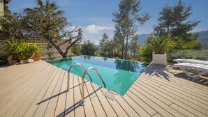 Une piscine à débordement dotée d’une terrasse en bois, entourée de plantes en pot et d’arbres, et offrant une vue imprenable sur un paysage montagneux.