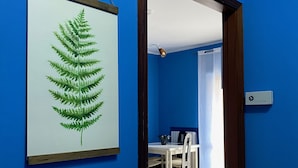 A room with a blue wall, a framed fern artwork, a dining area with a table and chairs, and a tiled floor.
