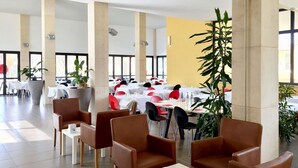 Un vaste espace intérieur aménagé en salle à manger, avec de hauts plafonds, de grandes fenêtres et un mur d’accent jaune. Les tables sont dressées avec des nappes blanches et des chaises rouges.
