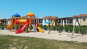 Un terrain de jeux avec des toboggans et des structures d’escalade, entouré de maisons dotées de balcons et de parasols.