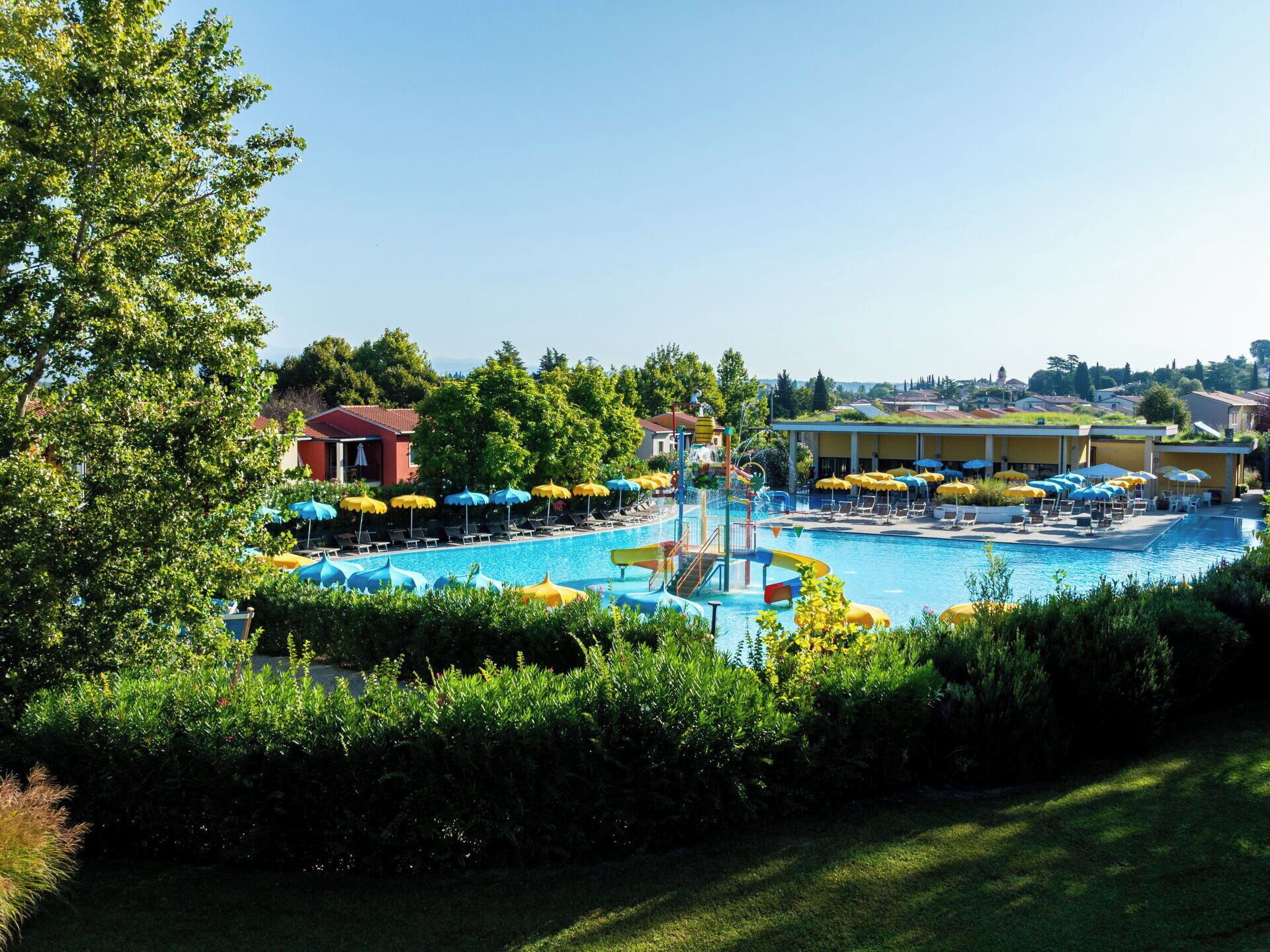 A swimming pool with a water slide, surrounded by lounge chairs and umbrellas, with buildings and trees in the background.