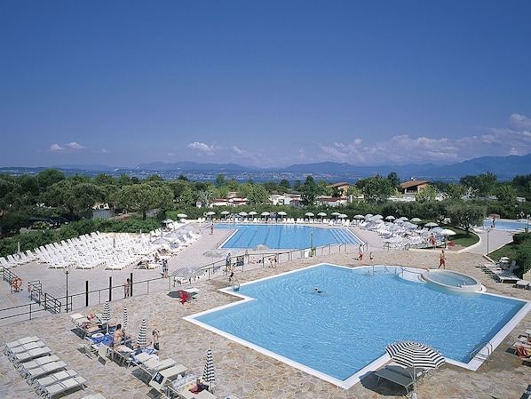 A large outdoor swimming pool with multiple lanes, surrounded by lounge chairs and umbrellas, set against a backdrop of mountains and a clear sky.