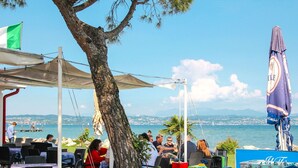 An outdoor seating area with tables and chairs, a large tree, and a view of the sea. - Beautiful apartment in villa for 4 people with A/C, WIFI, pool, TV and terrace (Sirmione)