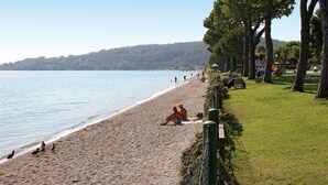 Ein Strand mit badenden Menschen und Schwimmern, eine begrünte Fläche mit Bäumen und ein ruhiges Meer.