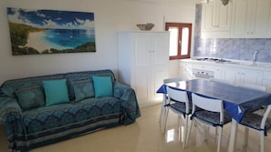 Un salon chaleureux avec un canapé et une table à manger, une cuisine aux meubles blancs et un tableau représentant une scène de plage accroché au mur.