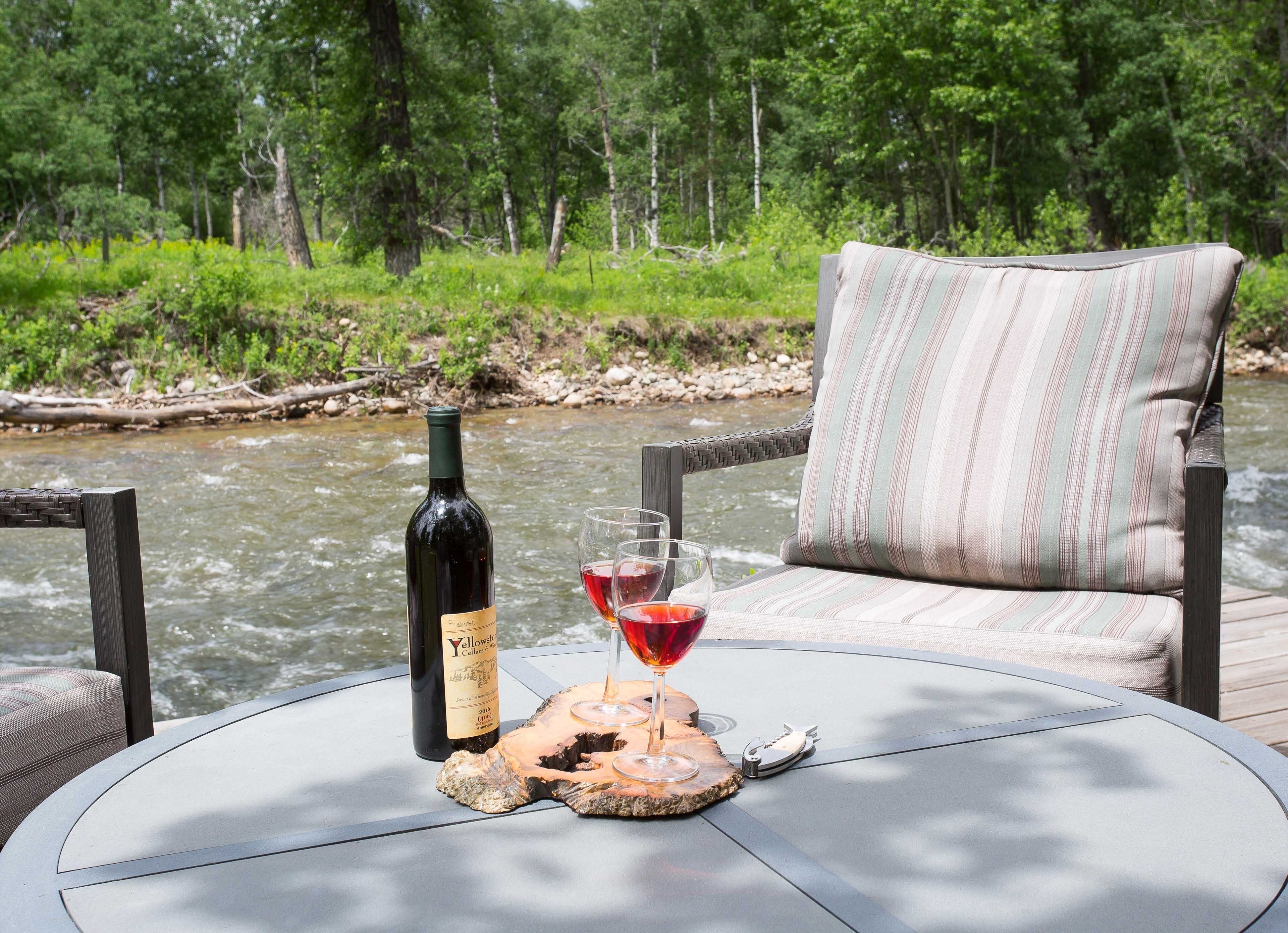 A table with a bottle of wine and a glass of red wine, a folded napkin, and a wooden coaster. The table is outdoors with a view of a river and trees in the background.