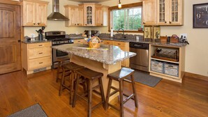 A kitchen with wooden cabinets, granite countertops, a central island with stools, stainless steel appliances, and a large window providing natural light. - Luxury Riverfront Beartooth Mountain Lodge - Private Retreat! Visit Yellowstone (Roscoe)