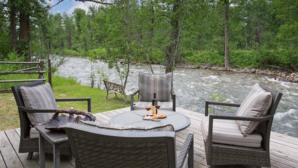 A wooden deck with wicker furniture and a view of a river. - Luxury Riverfront Beartooth Mountain Lodge - Private Retreat! Visit Yellowstone (Roscoe)