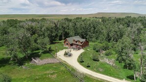 A large, two-story log cabin with a wrap-around porch, surrounded by dense forest and a dirt road leading to it. - Luxury Riverfront Beartooth Mountain Lodge - Private Retreat! Visit Yellowstone (Roscoe)