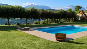 Una piscina con una silla de madera y un banco, rodeada de césped cuidado y árboles, con vista a la montaña al fondo.
