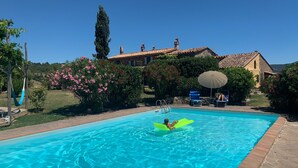 Une piscine avec une personne sur un flotteur, entourée d’un jardin et d’une maison avec un toit en tuiles.