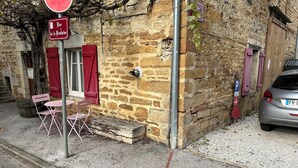 Un edificio de piedra con una ventana con persianas rojas, una pequeña zona de estar al aire libre con sillas rosadas y un coche aparcado.