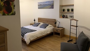 Un dormitorio con cama de madera, mesitas de noche, un escritorio y una silla. Hay un cuadro en la pared y una estantería con libros y plantas.
