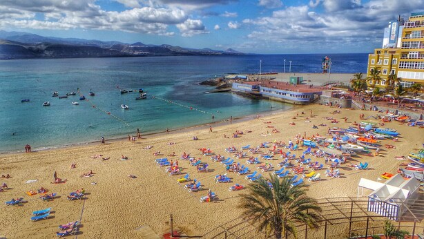 A beach with numerous sun loungers and umbrellas, a pier with boats, and a mountain range in the background. - Stratus Loft Gran Canaria, elegance on Las Canteras beach. (Las Palmas de Gran Canaria)
