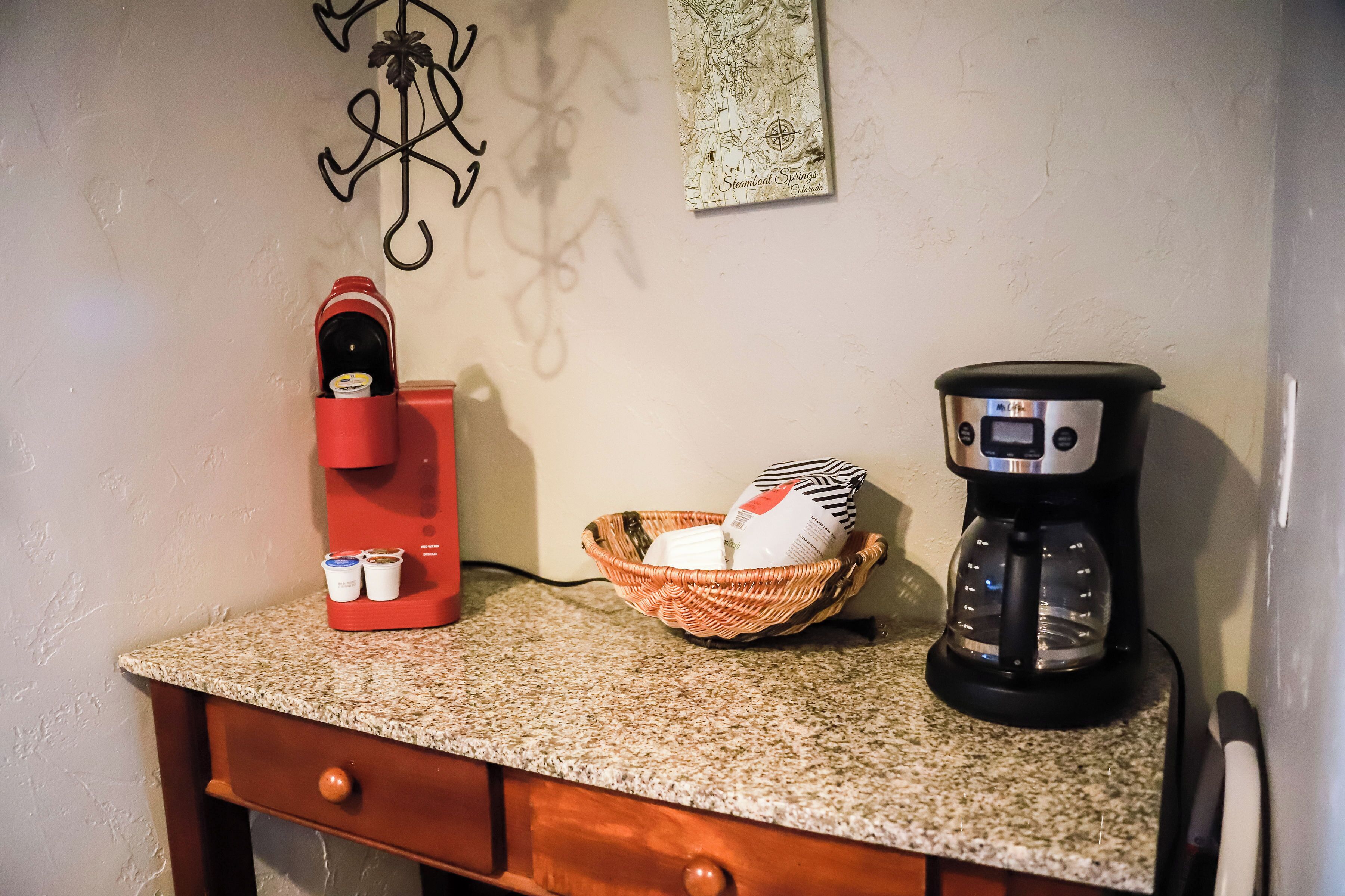 Un piano di lavoro della cucina con una macchina da caffè, un elettrodomestico rosso e un cesto con una bottiglia e un sacchetto.