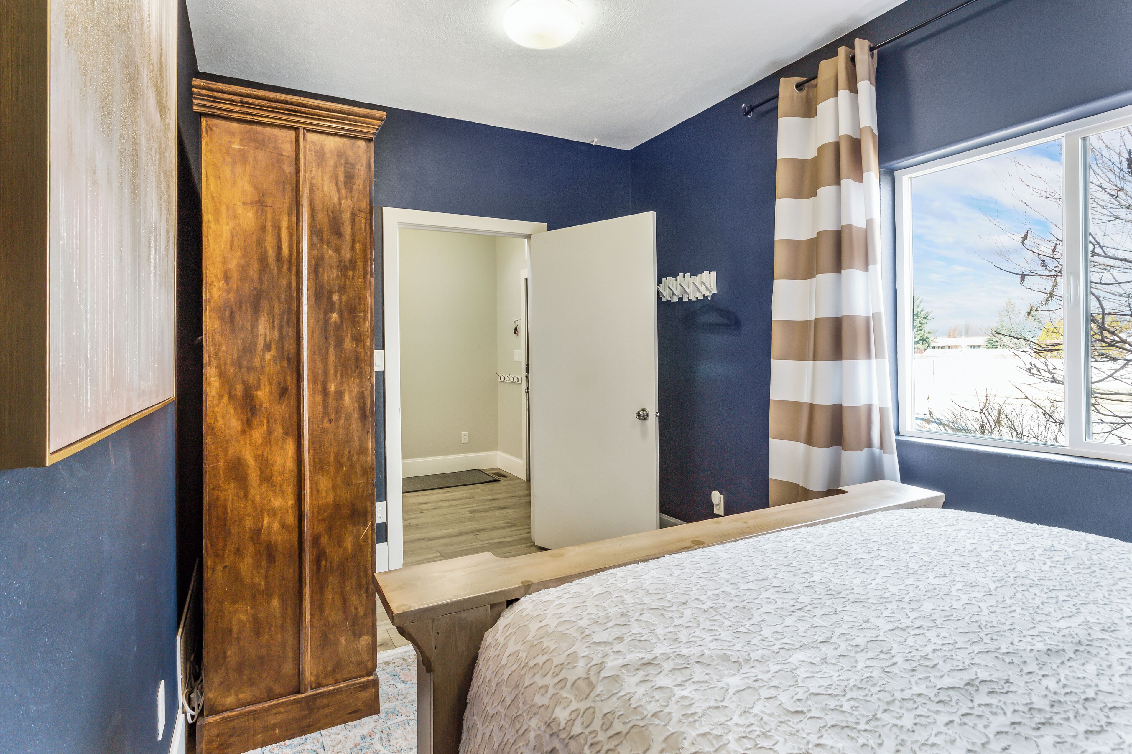 A bedroom with a bed, a wooden wardrobe, a door leading to a bathroom, and a window with curtains.